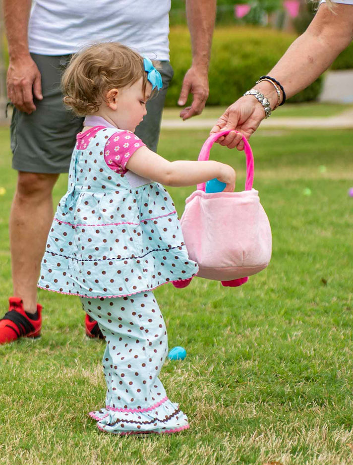 A little girl dressed in bright spring colors holds up her Easter basket while her older companion drops a colorful egg in
