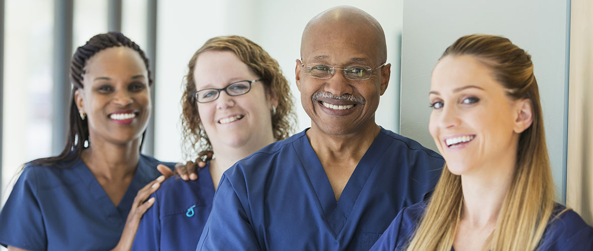 Three female nurses and a male nurse wearing a stethoscope stand outside an emergency room entrance.