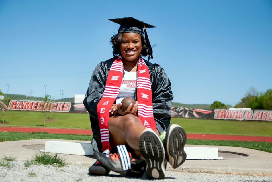 Danielle Moss, in her cap and gown and holding a shot put, sits on the track where she competed during her college years.